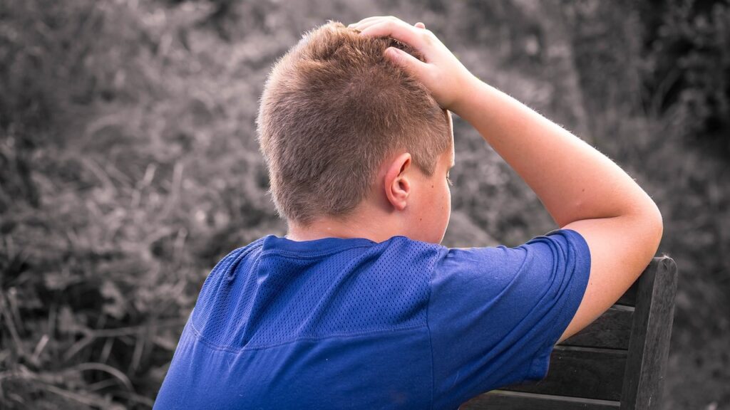 A white boy with blond hair and a blue shirt sits on a bench with his elbows propped up on the back of the bench, and his head in his hands.