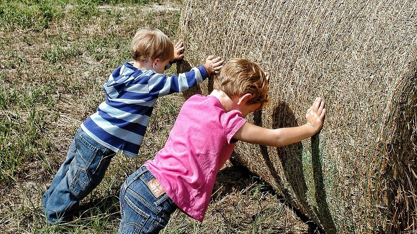 Two boys push against a huge bail of hay in a farm field on a sunny day.