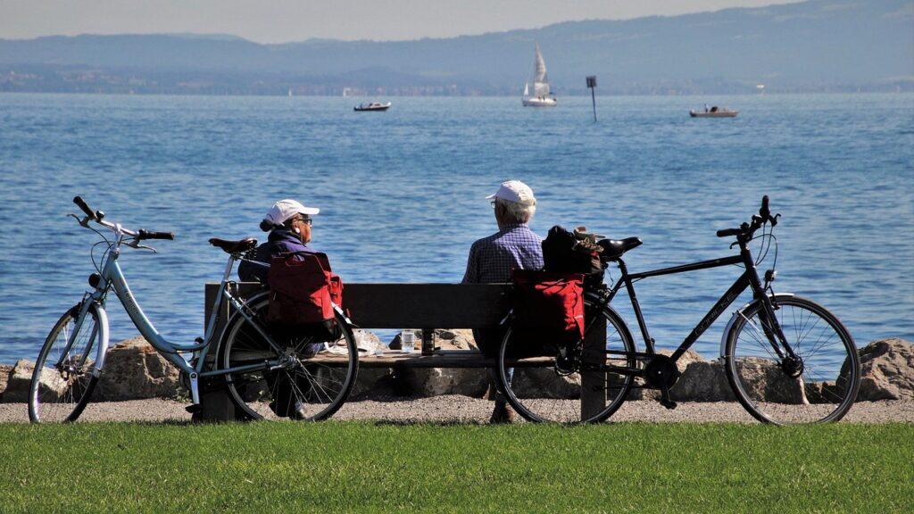 A woman and man sit on a park bench, looking out at a body of water and mountains beyond it. There are two bicycles next to them on the two ends of the bench.